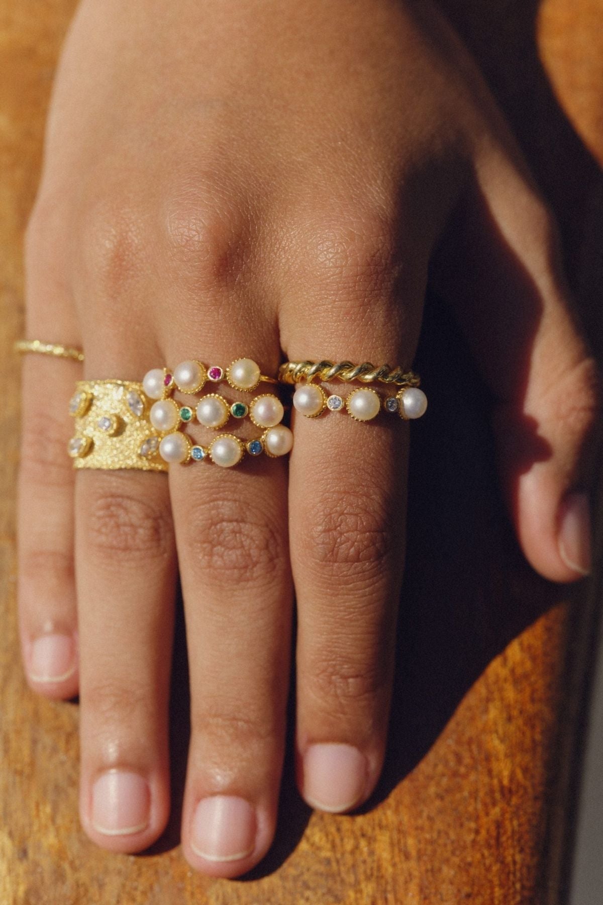 Hand wearing multiple gold and pearl rings on a wooden surface