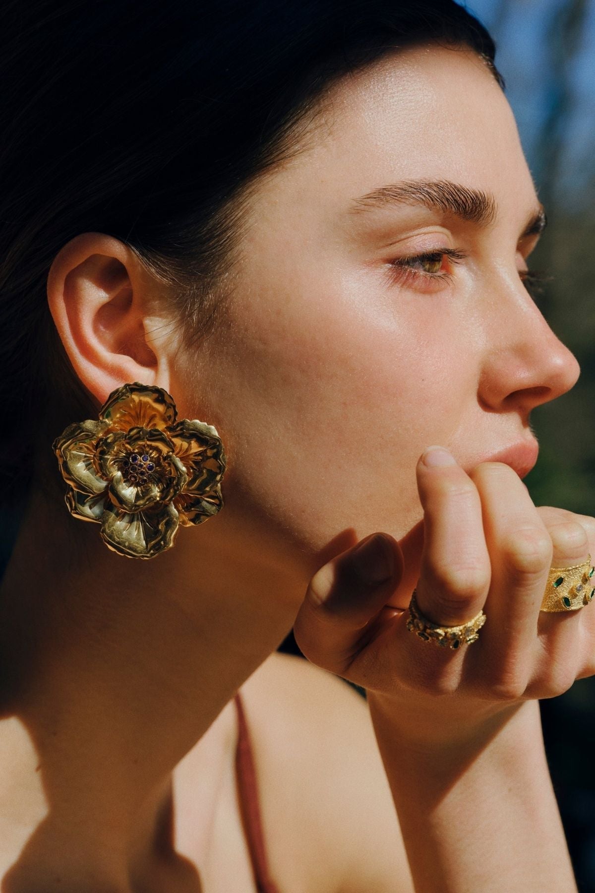 Close-up of a person wearing gold floral earrings and rings with a blurred natural background