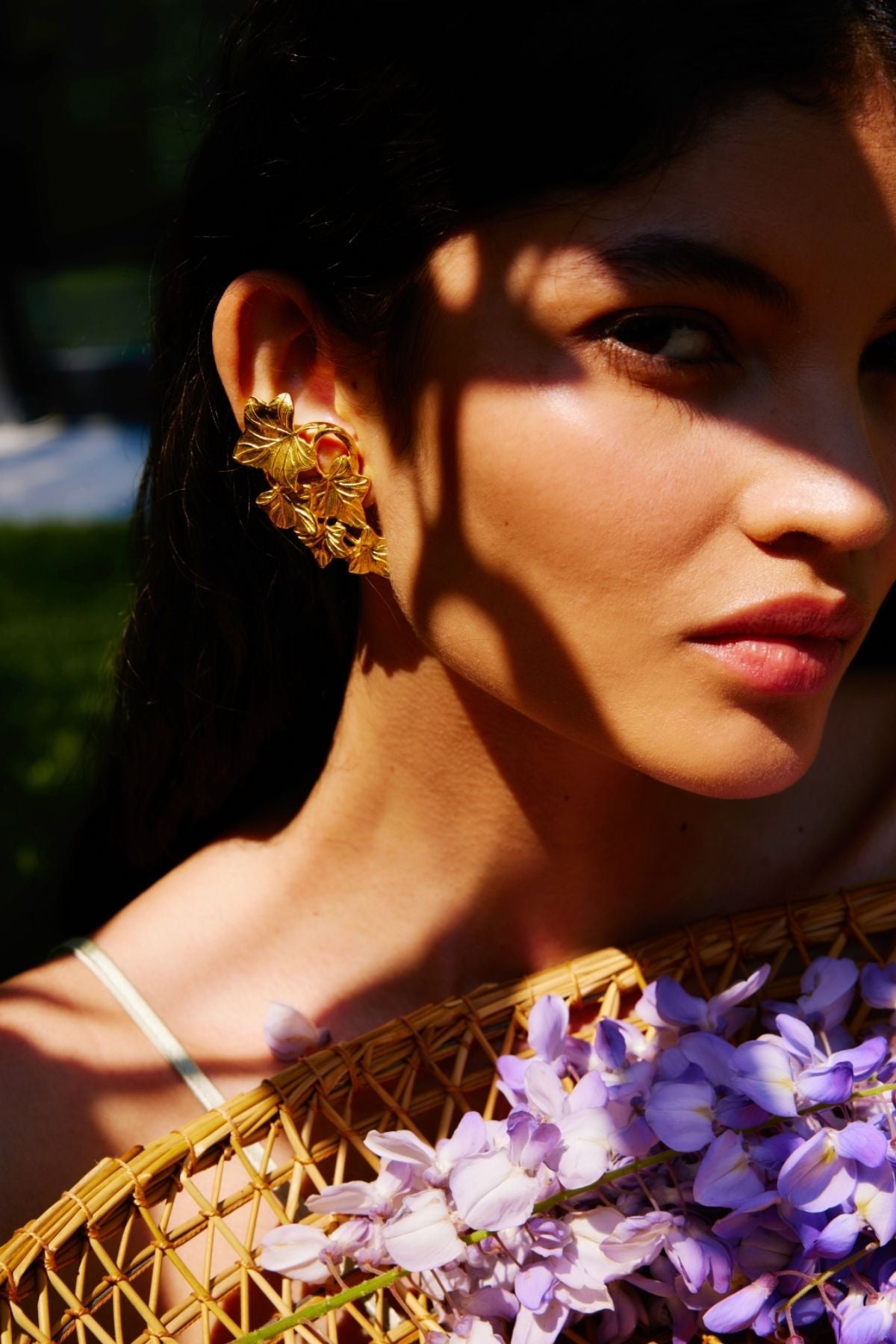 Woman wearing gold earrings with a basket of purple flowers, sunlit outdoors.