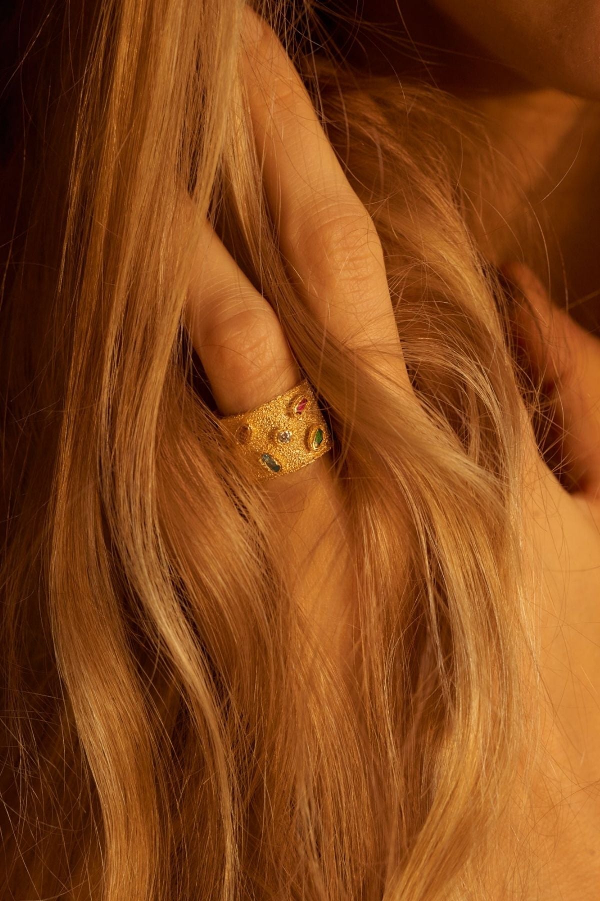 Close-up of a hand with a gold ring featuring green gemstones, held against wavy blonde hair.
