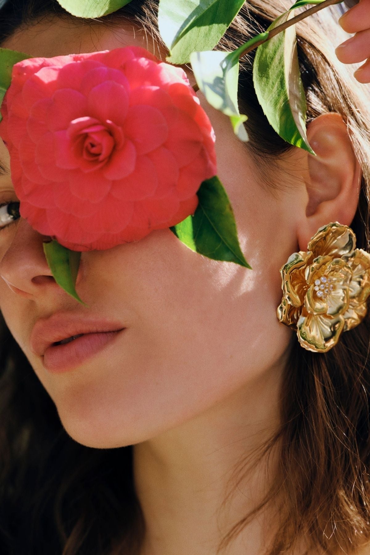 Close-up of a woman's face with a pink flower covering one eye and a gold floral earring.