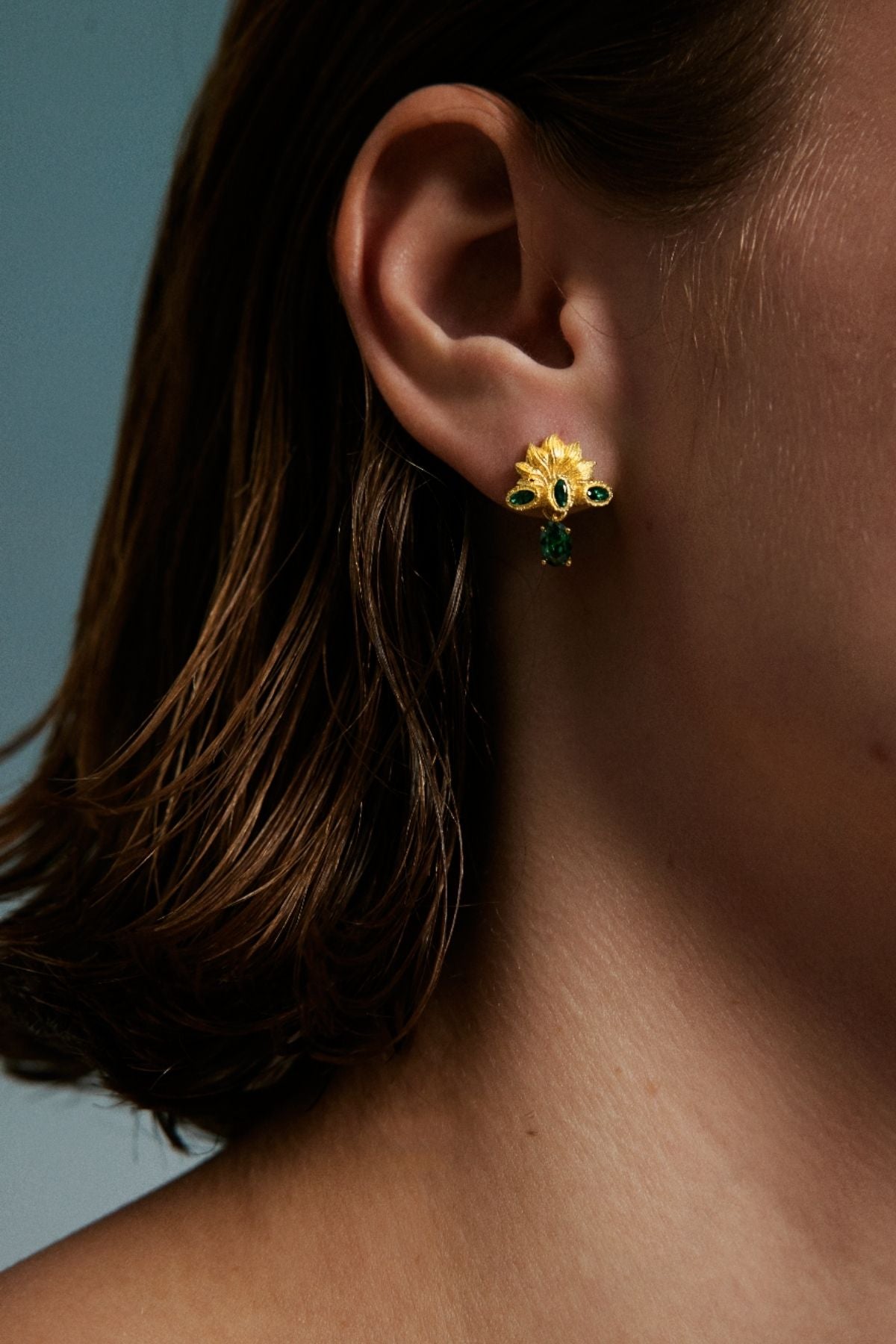 Close-up of an ear wearing a gold earring with green gemstones against a blurred background