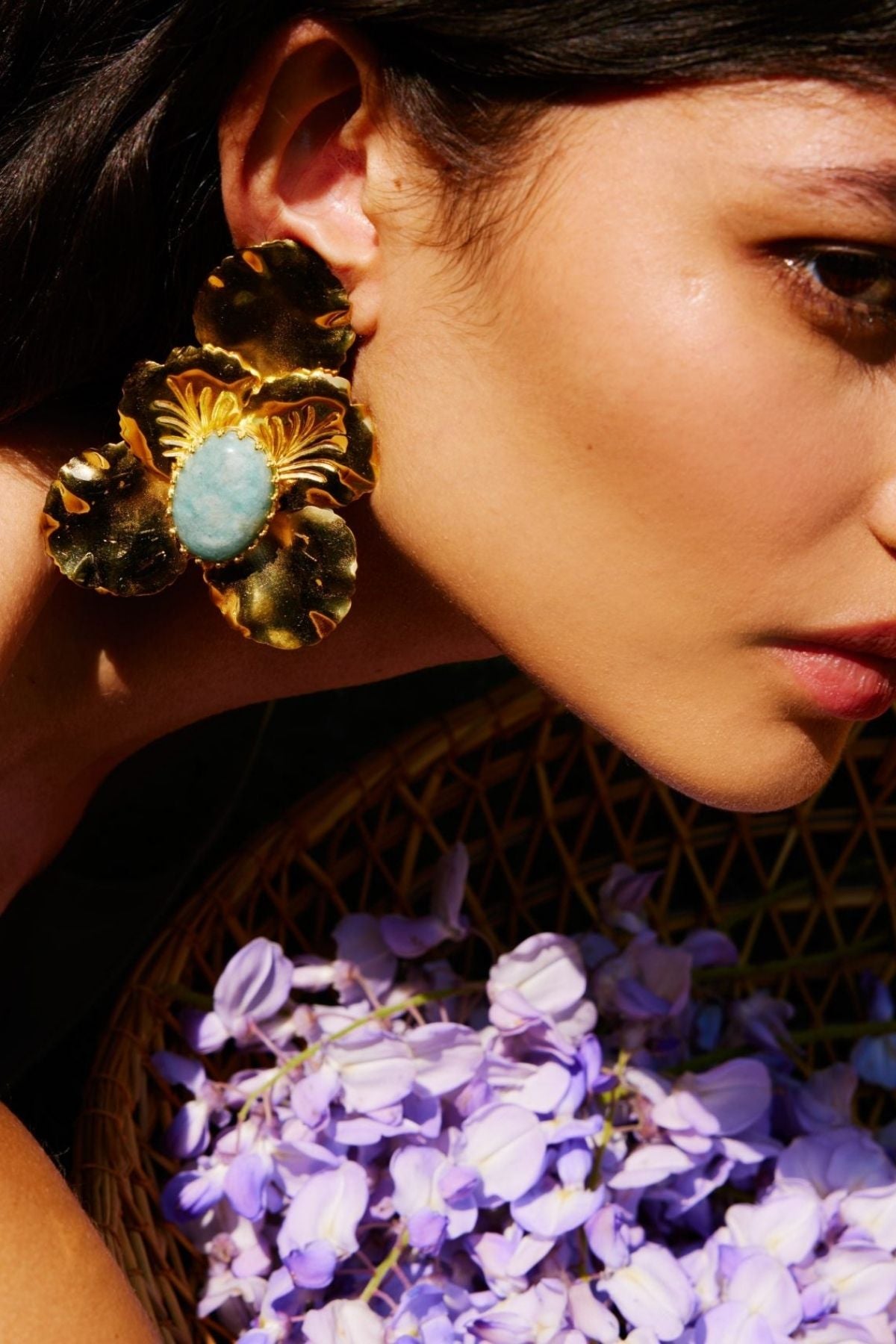 Close-up of a person wearing gold earrings with a blue stone, holding purple flowers.